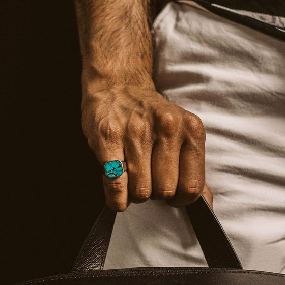 Un homme en pantalon blanc, tenant un sac en cuir et portant une bague de sceau pour homme « Nuri » turquoise de la marque Ninetwofive.