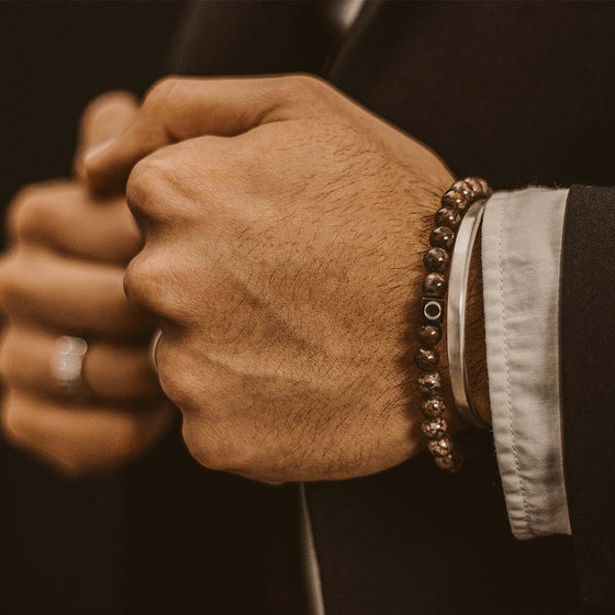 Een man in een zwart pak draagt een bruine kralenarmband van Albuna en een manchetband van Ninetwofive.