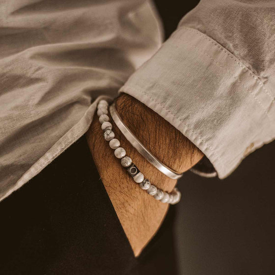 Model in a white shirt wearing the Ninetwofive Alrukham mens white beaded bracelet and a silver cuff, with his hand in his black trouser pocket.