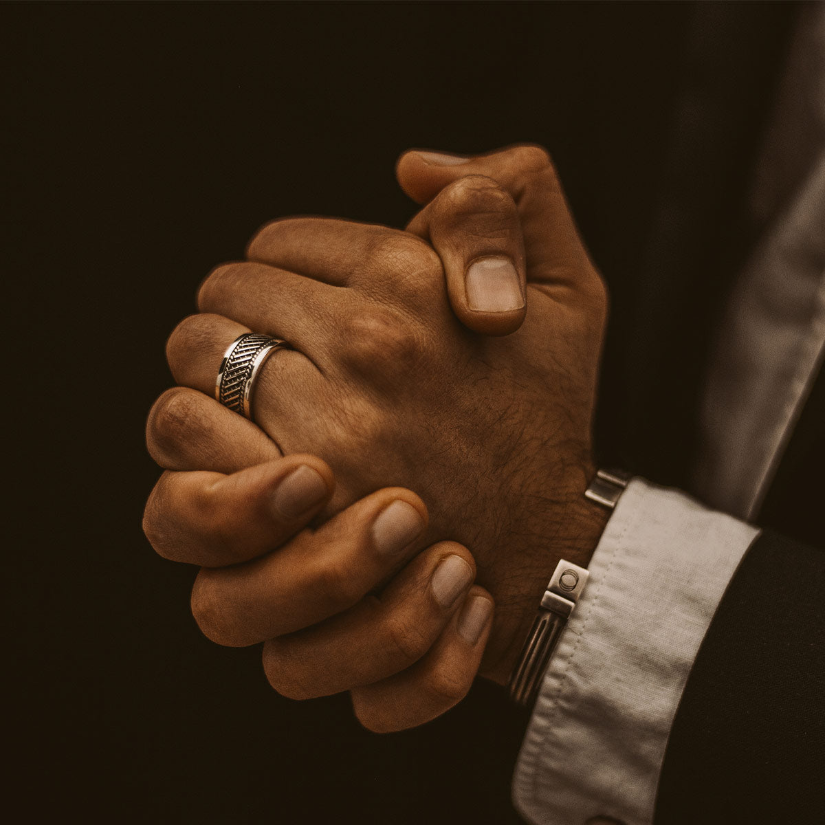 Een man in pak houdt zijn handen bij elkaar terwijl hij een Kaliq - Geoxideerde Sterling Zilveren Ring 10mm draagt.