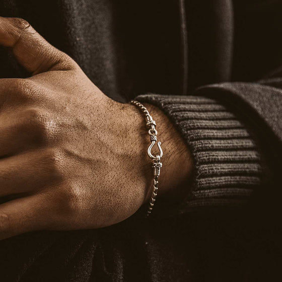 Die Hand eines Mannes in einem kuscheligen schwarzen Pullover, die das minimalistische Herrenarmband „Ninetwofive“ von Emir aus Sterlingsilber hält.