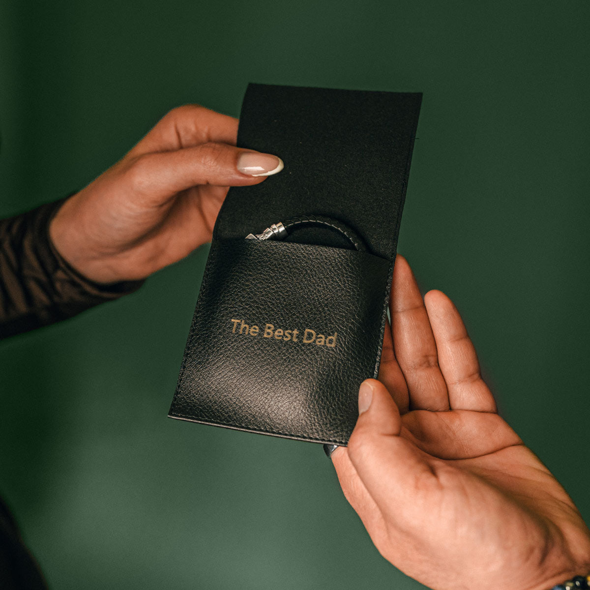 Pochette en cuir noir portant l'inscription « The Best Dad », tenue par deux mains sur fond vert