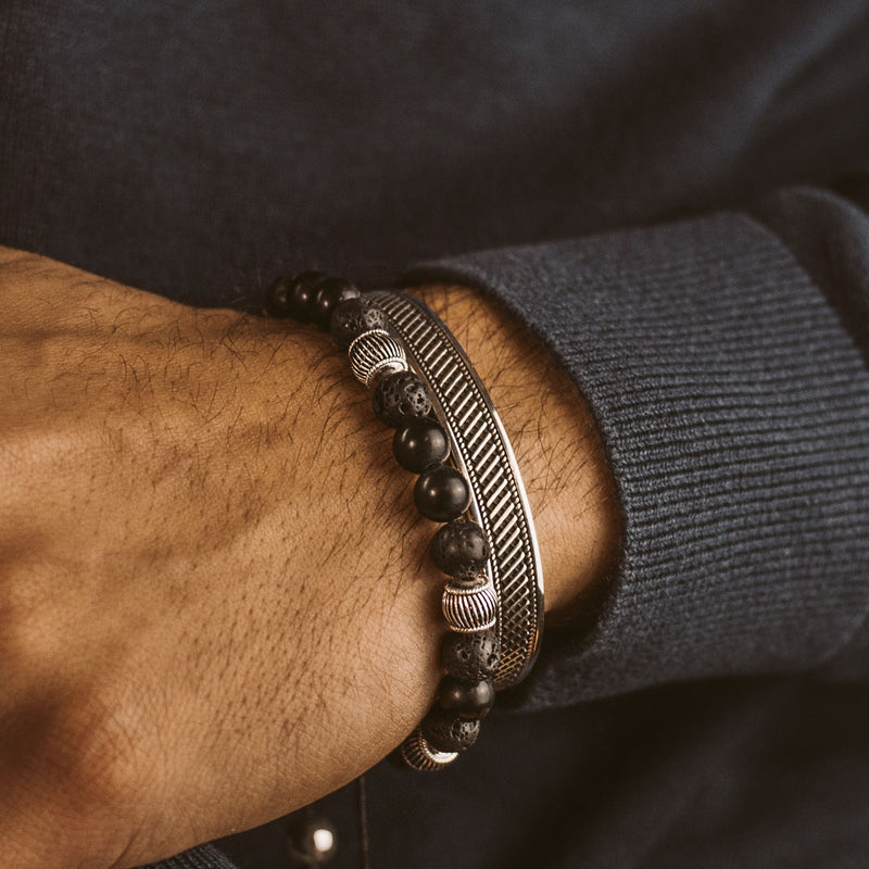 A man in blue sweater wears a Kaliq adjustable onyx black beaded mens bracelet and silver cuff. 