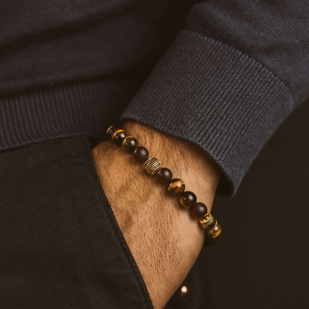 Een man draagt een Kaliq - Verstelbare Tijgeroog Kralenarmband in Goud 8mm en ebbenhouten stenen armband.