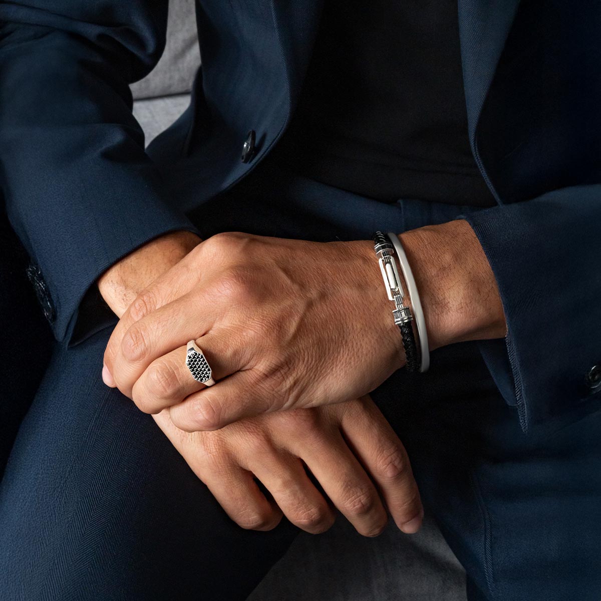 Close-up of a person wearing a suit with hands clasped together, showing a ring and bracelets from ninetwofive