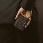 Een man in een zwart pak draait zich om en toont de zwarte leren herenportemonnee van Ninetwofive . 