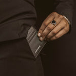 Man stopt een zwart leren minimalistische herenportemonnee in zijn zwarte broekzak en draagt de Ninetwofive zegelring.