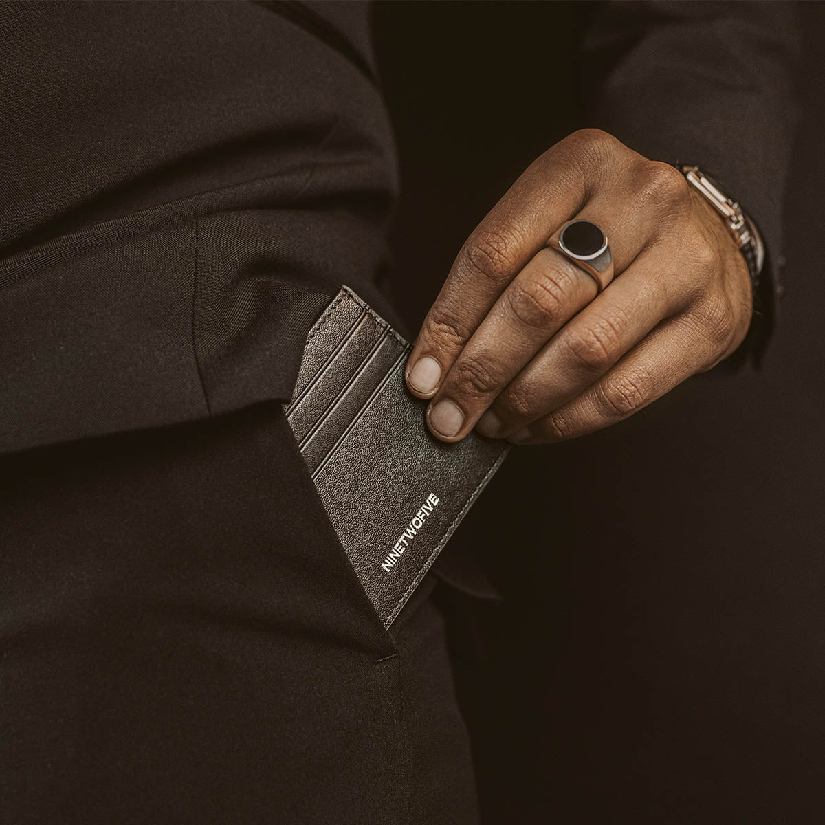 Man stopt een zwart leren minimalistische herenportemonnee in zijn zwarte broekzak en draagt de Ninetwofive zegelring.