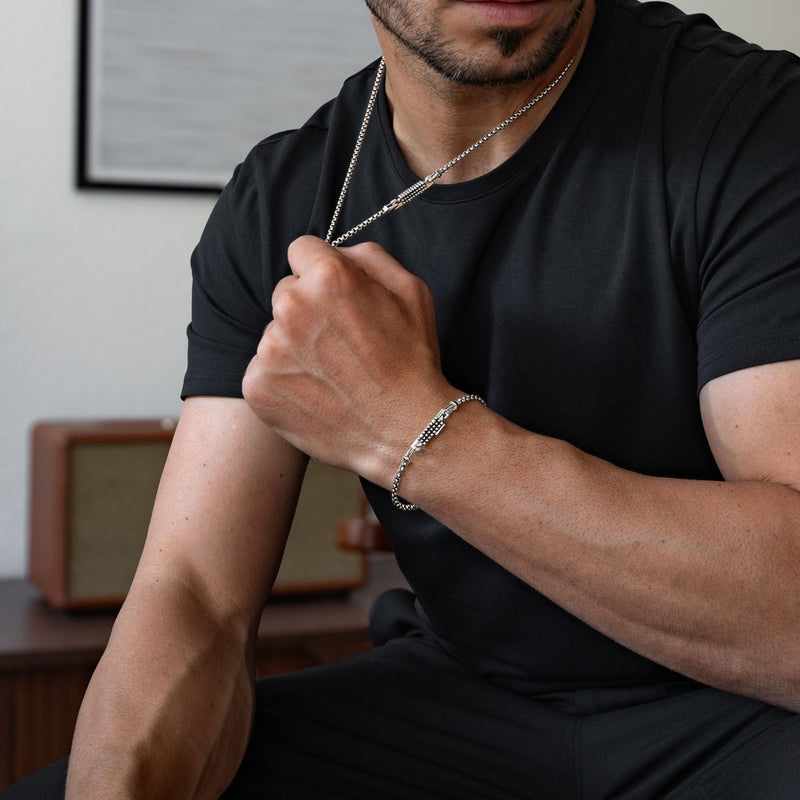 Ein Mann, der ein schwarzes T-Shirt und ein silbernes Ankerkettenarmband trägt und in einem Raum vor einem neutralen Hintergrund sitzt