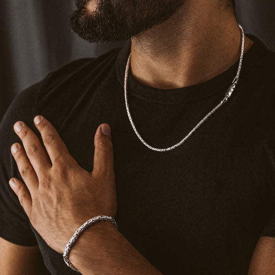 A man in beard wearing black t shirt is showing Turath sterling silver byzantine mens bracelet and chain.