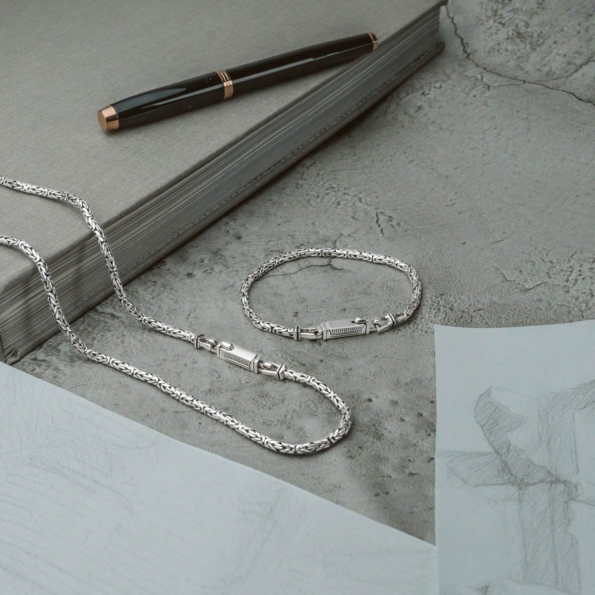 Ramsi men’s sterling silver bracelet and chain from Ninetwofive placed on a desk with sketch paper, pen and grey book at the side.