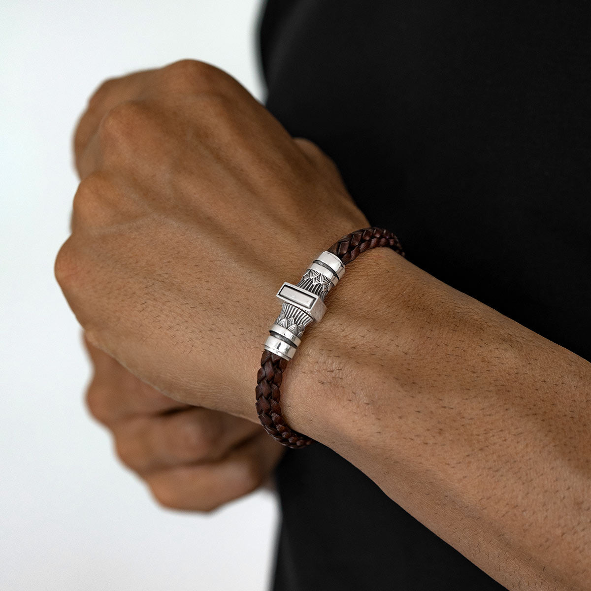 A close-up of a man's wrist wearing a dark brown braided leather bracelet with a silver-toned metal clasp.