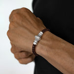 A close-up of a man's wrist wearing a dark brown braided leather bracelet with a silver-toned metal clasp.