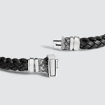 A black braided leather bracelet with a polished silver-toned rectangular clasp featuring engraved geometric patterns, displayed on a light gray background.