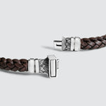 A close-up of a dark brown braided leather bracelet with a silver-toned rectangular clasp featuring engraved leaf-like patterns, shown on a light gray background.