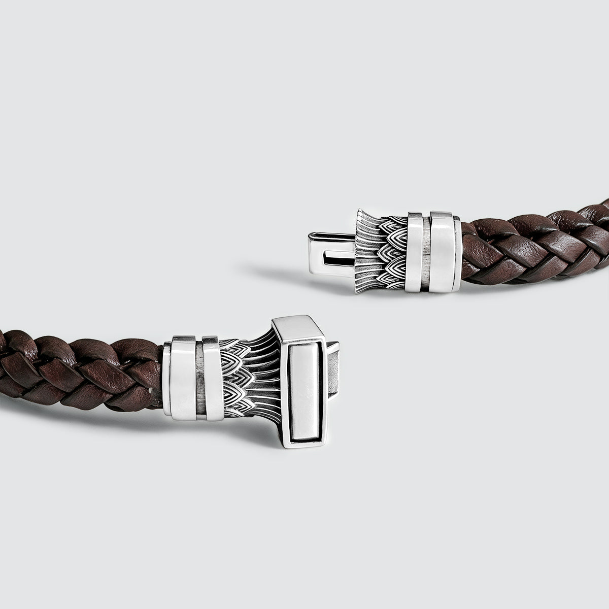A close-up of a dark brown braided leather bracelet with a silver-toned rectangular clasp featuring engraved leaf-like patterns, shown on a light gray background.