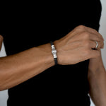 A close-up of a man's wrist wearing a black braided cord bracelet with a silver-toned rectangular clasp. The man is wearing a black shirt, and a silver band is visible on his other hand.