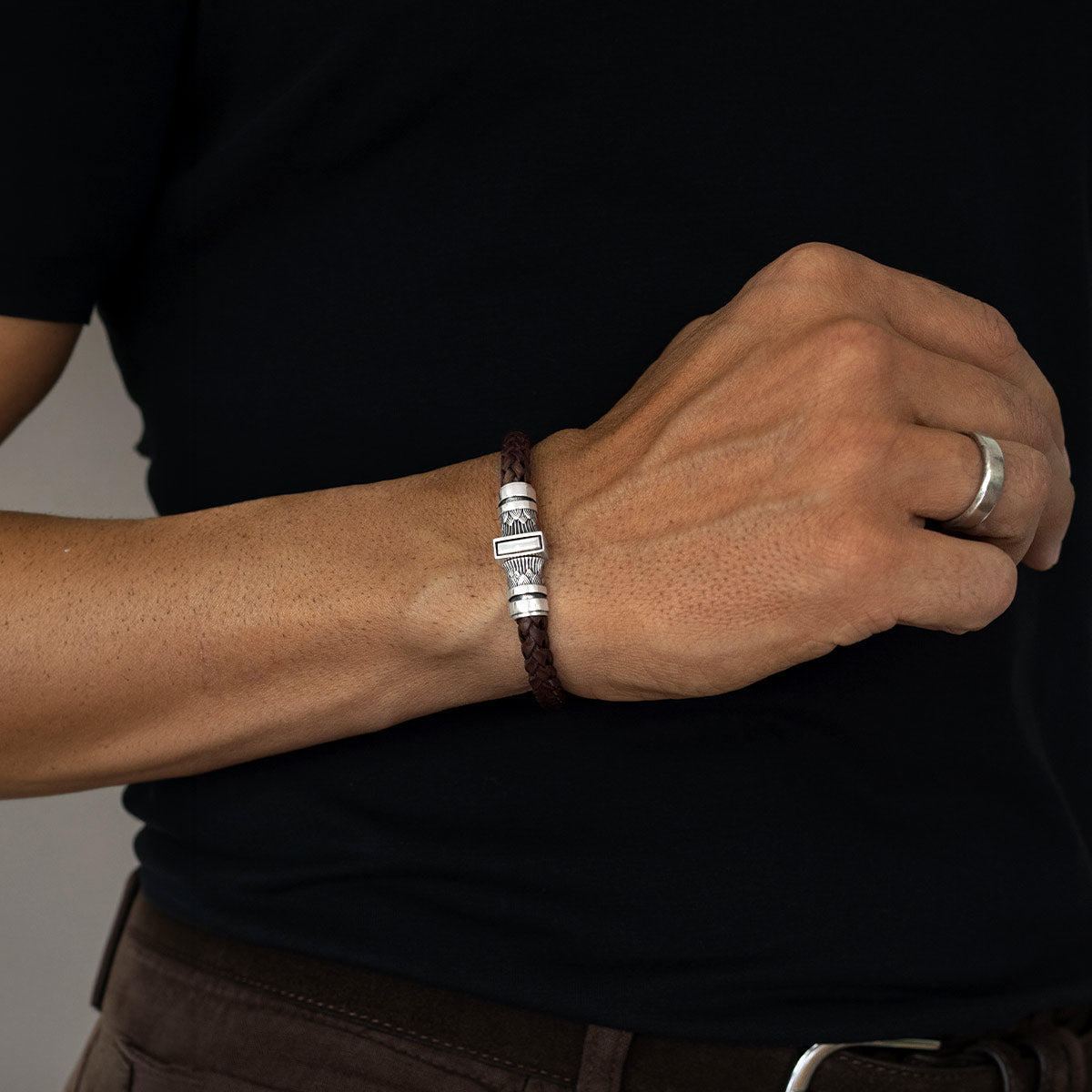 A close-up of a person's wrist wearing a dark brown braided leather bracelet with a silver-toned rectangular charm, paired with a silver ring on another finger, set against a black shirt.