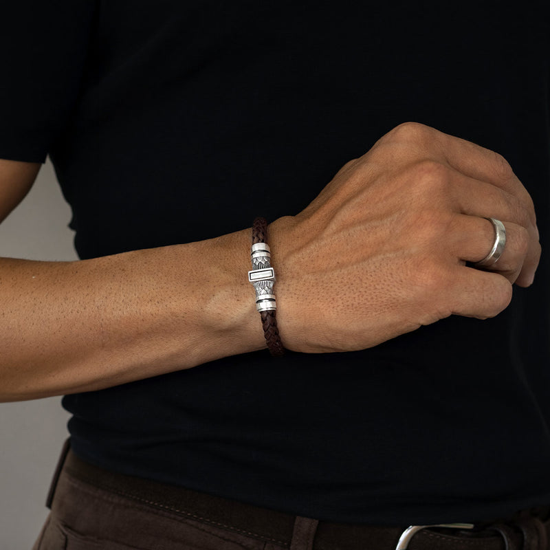 A close-up of a person's wrist wearing a dark brown braided leather bracelet with a silver-toned rectangular charm, paired with a silver ring on another finger, set against a black shirt.
