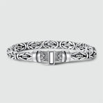 Ein silberfarbenes Metallarmband mit einem aufwendig strukturierten Muster und einem rechteckigen Verschluss in der Mitte, abgebildet vor einem hellgrauen Hintergrund.