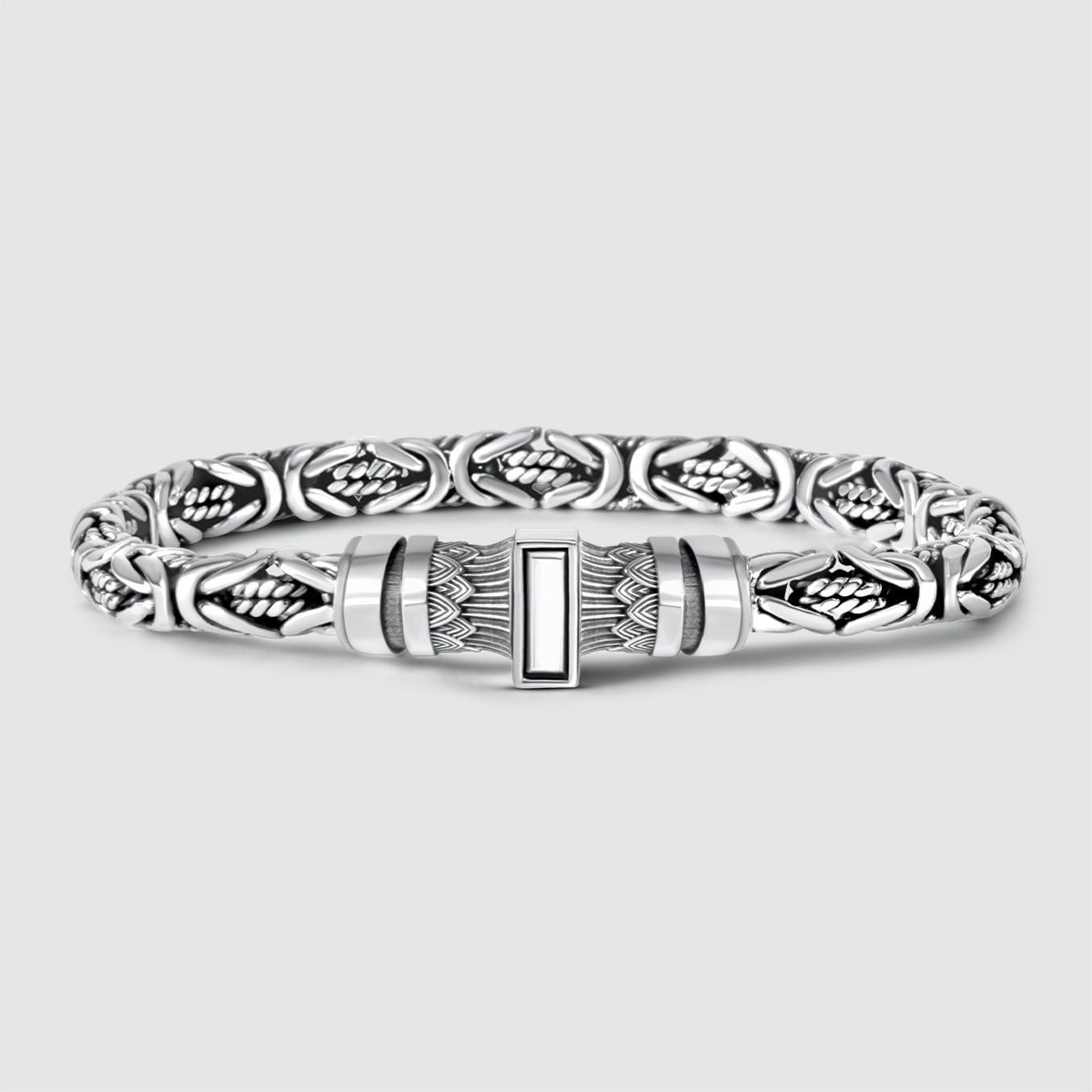 Ein silberfarbenes Metallarmband mit einem aufwendig strukturierten Muster und einem rechteckigen Verschluss in der Mitte, abgebildet vor einem hellgrauen Hintergrund.