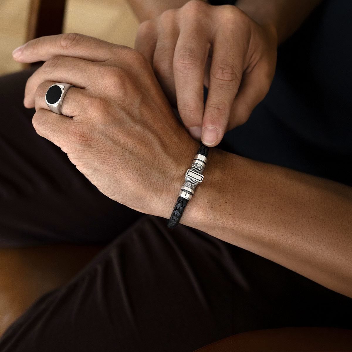 A close-up of a man's wrist wearing a black braided cord bracelet with silver-toned rectangular metal accents, while his other hand adjusts the bracelet. He is also wearing a silver signet ring with a black oval face.