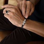 A close-up of a man's wrist wearing a black braided cord bracelet with silver-toned rectangular metal accents, while his other hand adjusts the bracelet. He is also wearing a silver signet ring with a black oval face.