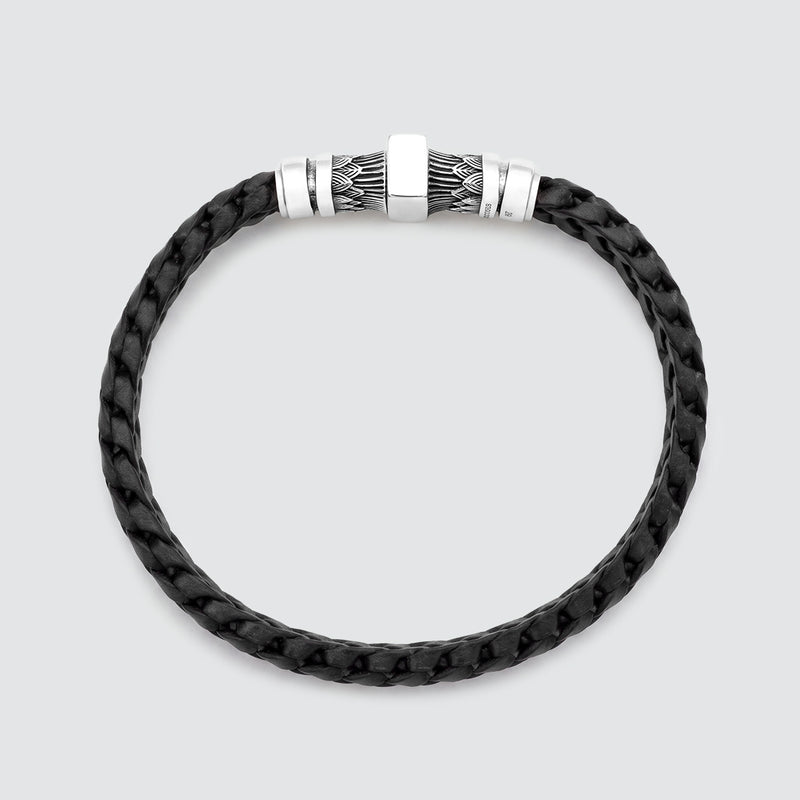Een zwarte, gevlochten leren armband met een zilverkleurige metalen sluiting, voorzien van een vierkant plaatje in het midden en gestructureerde details aan de zijkanten, afgebeeld tegen een lichtgrijze achtergrond.