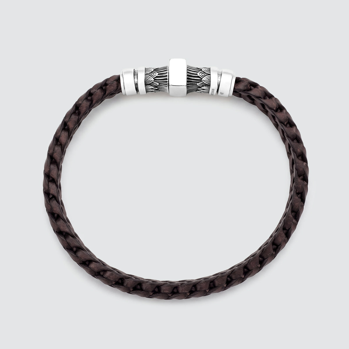 A dark brown braided leather bracelet with a silver-toned metal clasp featuring a textured geometric pattern, shown on a light gray background.
