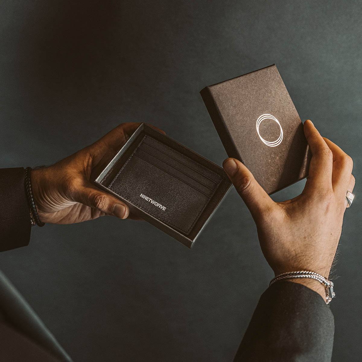 Model in een zwart pak opent de box Ninetwofive en onthult de echt zwart lederen minimalistische herenportemonnee.