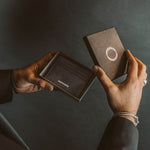 Model in een zwart pak opent de box Ninetwofive en onthult de echt zwart lederen minimalistische herenportemonnee.