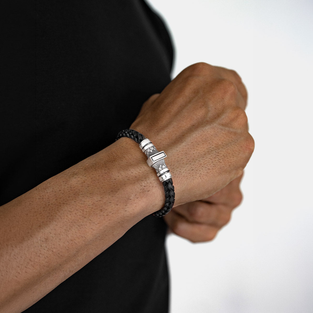 A close-up of a person's wrist wearing a black braided cord bracelet with polished silver-toned metal accents and a rectangular charm, against a plain light background.