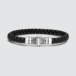 A black braided leather bracelet with a polished silver-toned metal clasp featuring geometric and feather-like engravings, set against a light gray background.