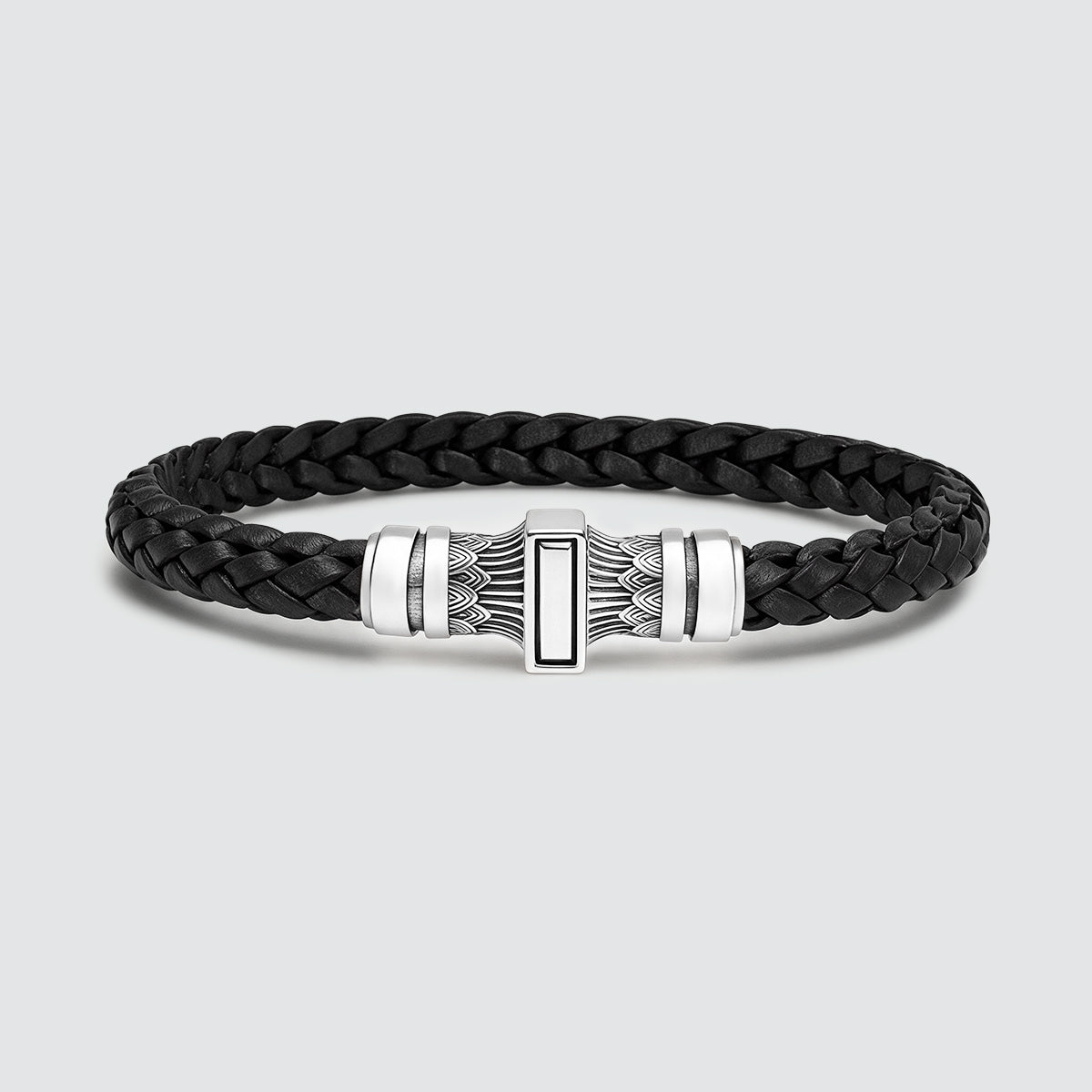 A black braided leather bracelet with a polished silver-toned metal clasp featuring geometric and feather-like engravings, set against a light gray background.