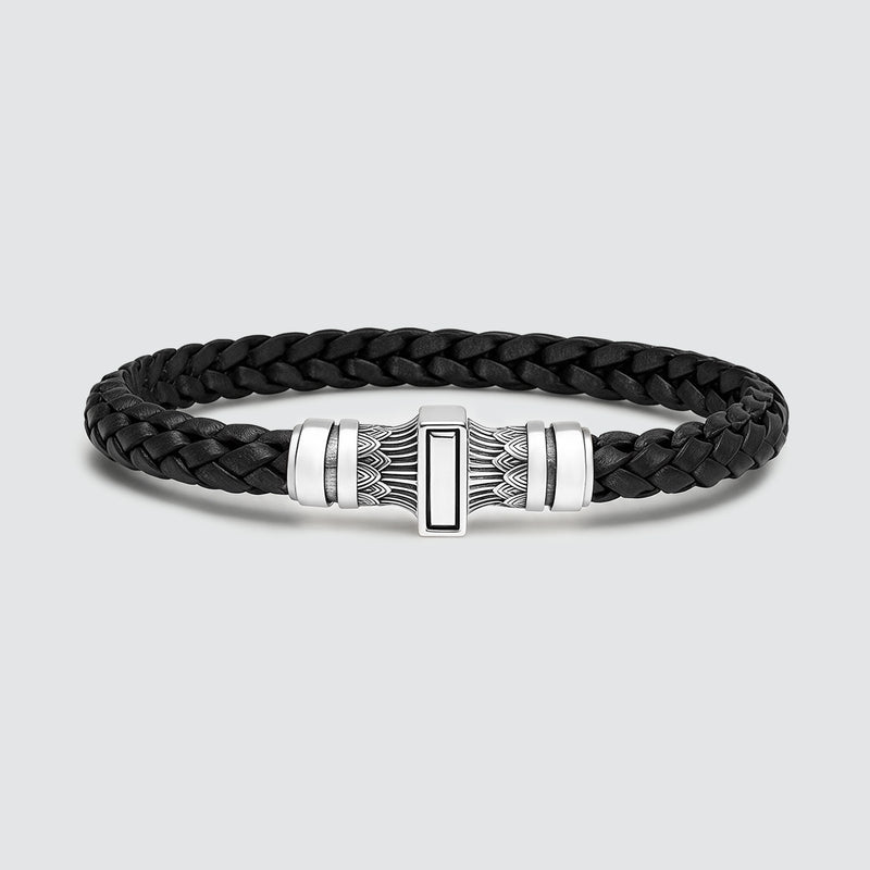 Een zwarte, gevlochten leren armband met een gepolijste, zilverkleurige metalen sluiting met geometrische en veerachtige gravures, set tegen een lichtgrijze achtergrond.