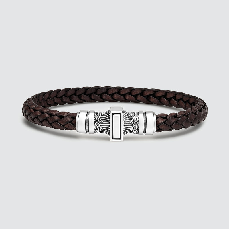 A dark brown braided leather bracelet with a silver-toned metal clasp featuring a central rectangular inlay, photographed on a light gray background.