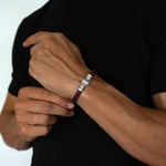 A close-up of a man's wrist wearing a dark brown braided cord bracelet with a silver-toned metal clasp and a small white stone accent.