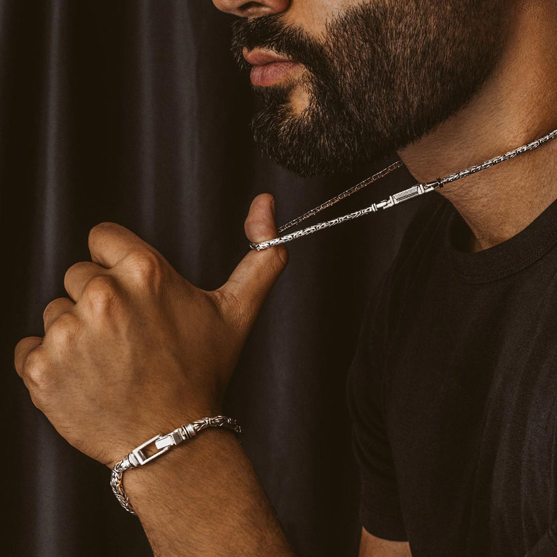 Een man met een baard laat de Turath Byzantine-ketting van sterlingzilver zien door deze van zijn duim af te trekken, en draagt een heren armband.