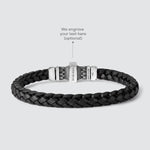 A dark brown braided leather bracelet with a silver-toned metal clasp, shown on a light gray background with an overlay pointing to the clasp and the text 'We engrave your text here'.