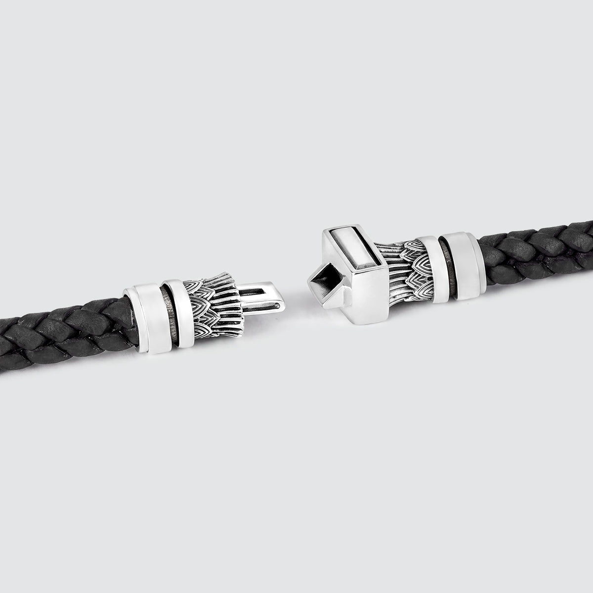 A close-up of a black braided leather bracelet with ornate silver-toned metal end caps, shown on a light gray background.
