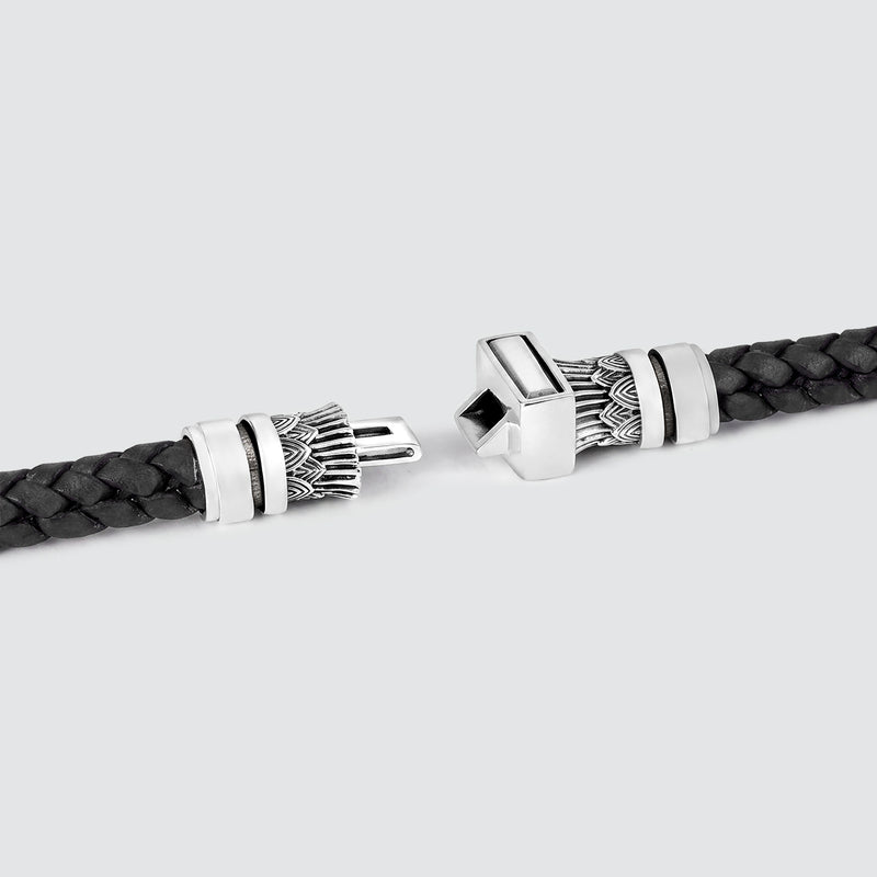 Een close-up van een zwarte, gevlochten leren armband met sierlijke zilverkleurige metalen eindstukken, afgebeeld tegen een lichtgrijze achtergrond.