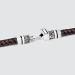 A close-up of a dark brown braided leather bracelet with ornate silver-toned metal clasps, shown against a light gray background.