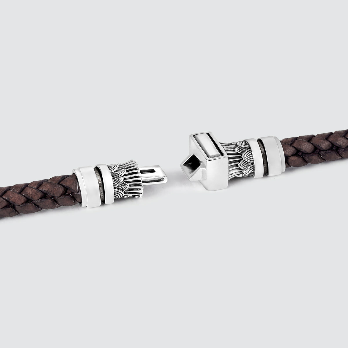 A close-up of a dark brown braided leather bracelet with ornate silver-toned metal clasps, shown against a light gray background.