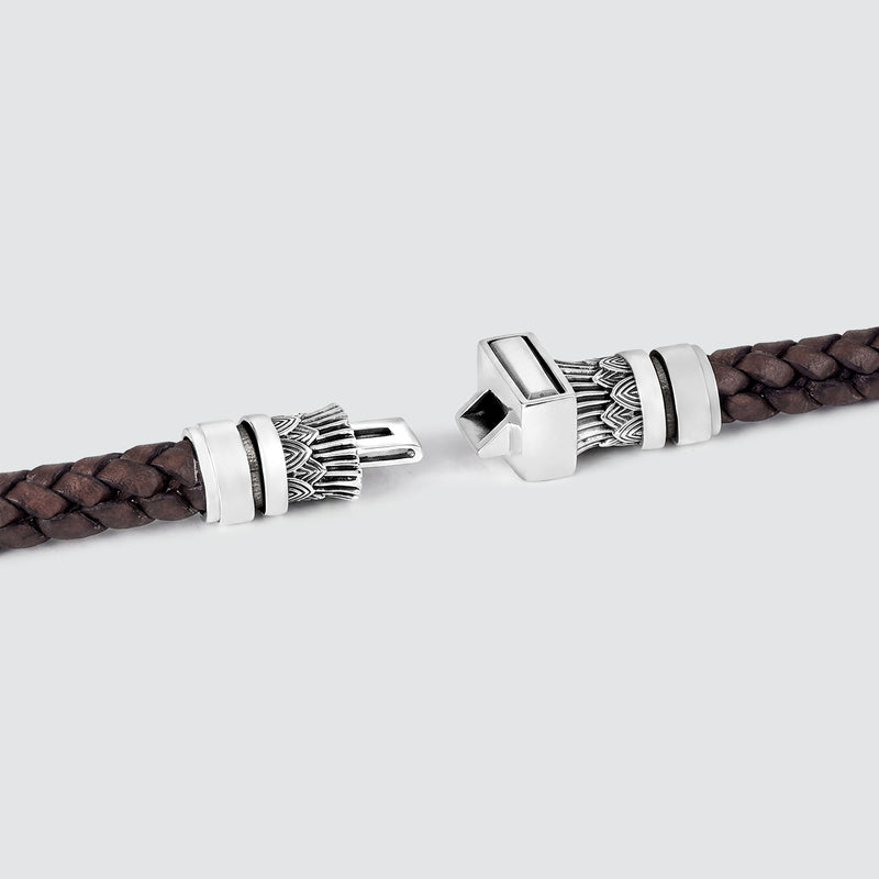 A close-up of a dark brown braided leather bracelet with ornate silver-toned metal clasps, shown against a light gray background.