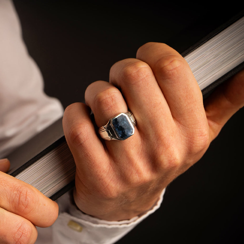 Un homme tient avec assurance un livre dans une main, tandis que l'autre main est ornée d'une magnifique bague Bariq - Blue Petersite Signet Ring 17mm.