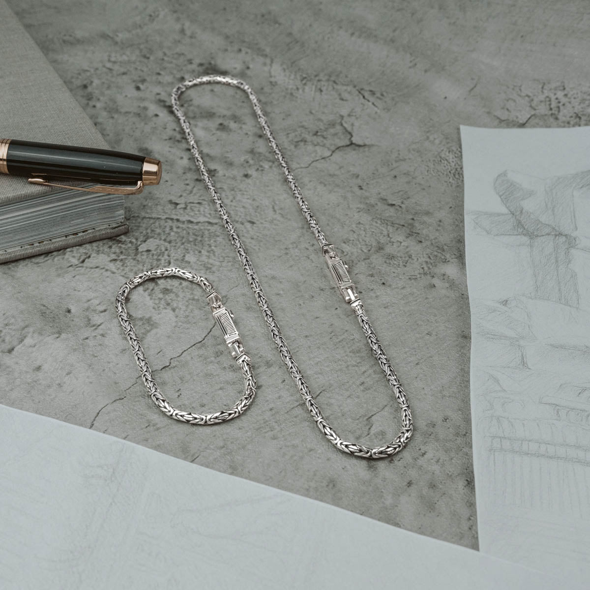Flatlay of Ramsi men’s sterling silver bracelet and chain from Ninetwofive with sketch paper, pen and grey book.