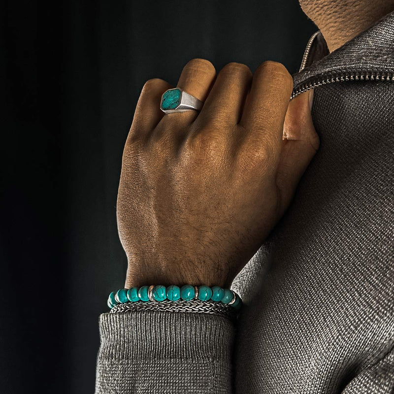 Hand van een man bij de rits van zijn trui, met de Nuri kralenarmband, een zegelring met turkoois steen en een sterling zilveren armband van Ninetwofive.