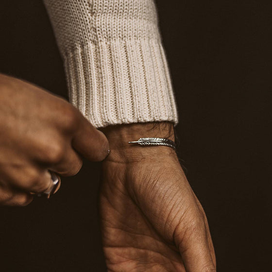 Un homme vêtu d'un pull confortable, portant un bracelet « Ninetwofive » de Zahir, fin massif, orné de plumes argentées, ainsi qu'une chevalière.