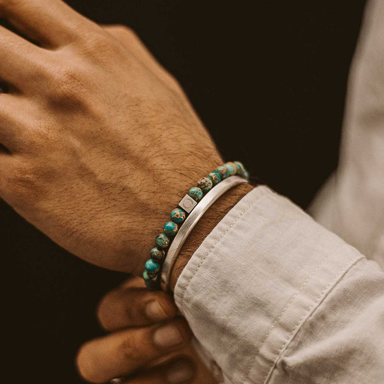 Een man in een wit overhemd draagt de Alfriuz-turquoise-kralenarmband voor heren, gecombineerd met een zilveren manchet van Ninetwofive.