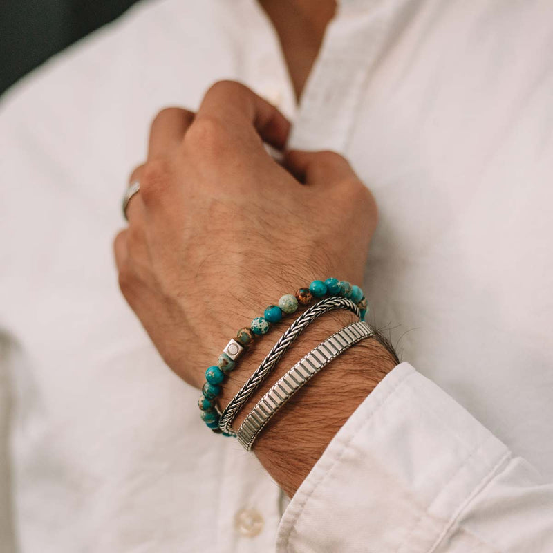 Een man in een wit overhemd die de ‘Ninetwofive’ van Alfiruz turquoise draagt: een kralenarmband voor heren en een manchet, terwijl hij de knoop van zijn overhemd vasthoudt.